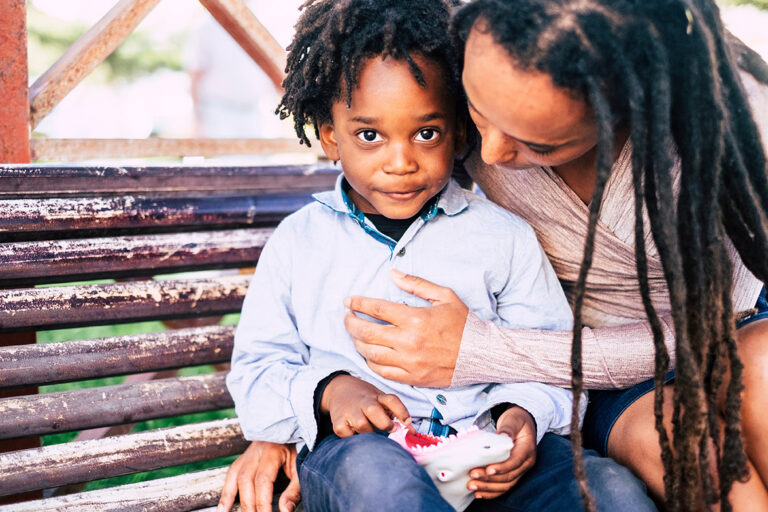 Small African American boy wearing blue long sleeve collard shirt looks calmly out from embrace of adult female seated next to him on park bench