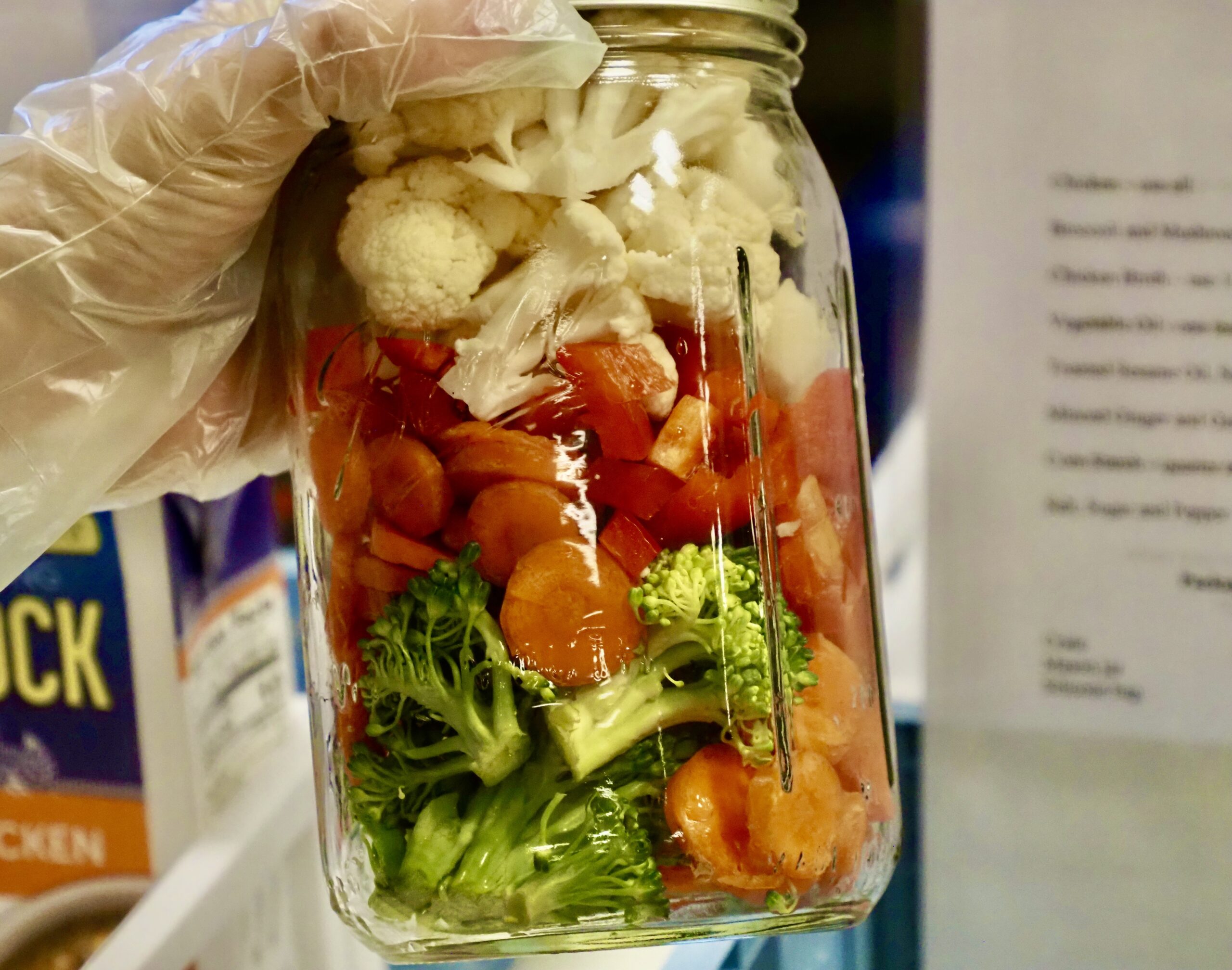 gloved hand holding a clear glass jar full of chopped broccoli, carrot and cauliflower.